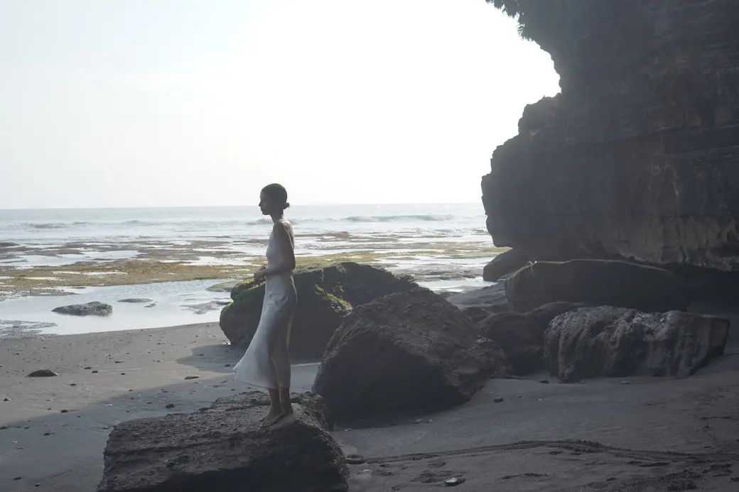 Woman in a light dress standing on a rock by the sea, backlit